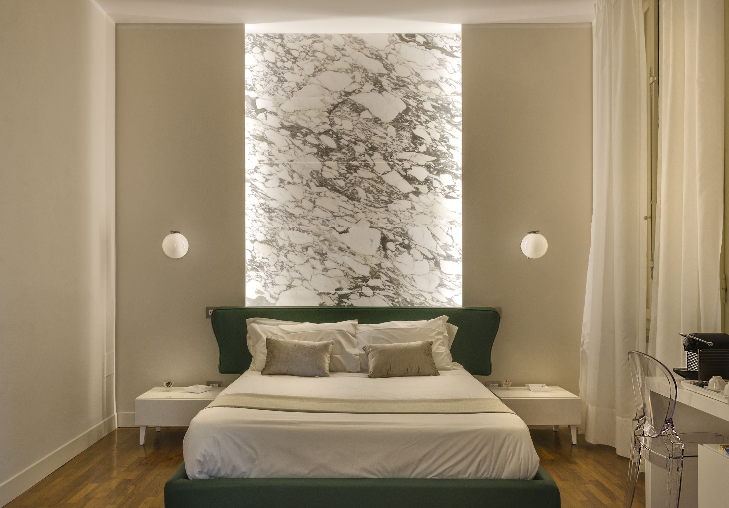 A modern bedroom with a large bed, green headboard, two bedside tables, and a marble-patterned wall.