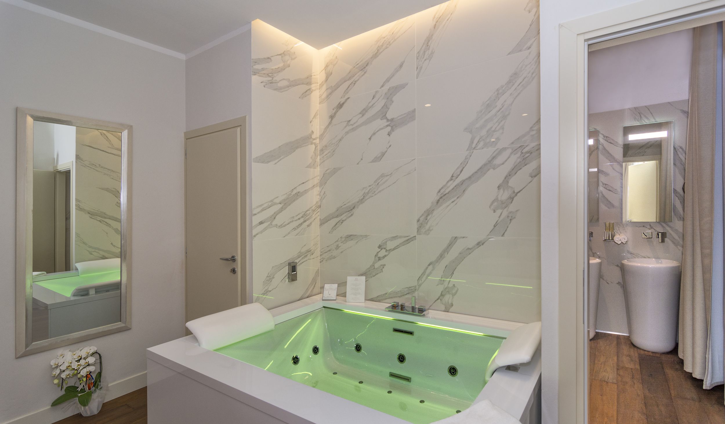 Een moderne badkamer met een marmeren wand, een grote spiegel, een groene whirlpool en een apart gelegen wastafel.