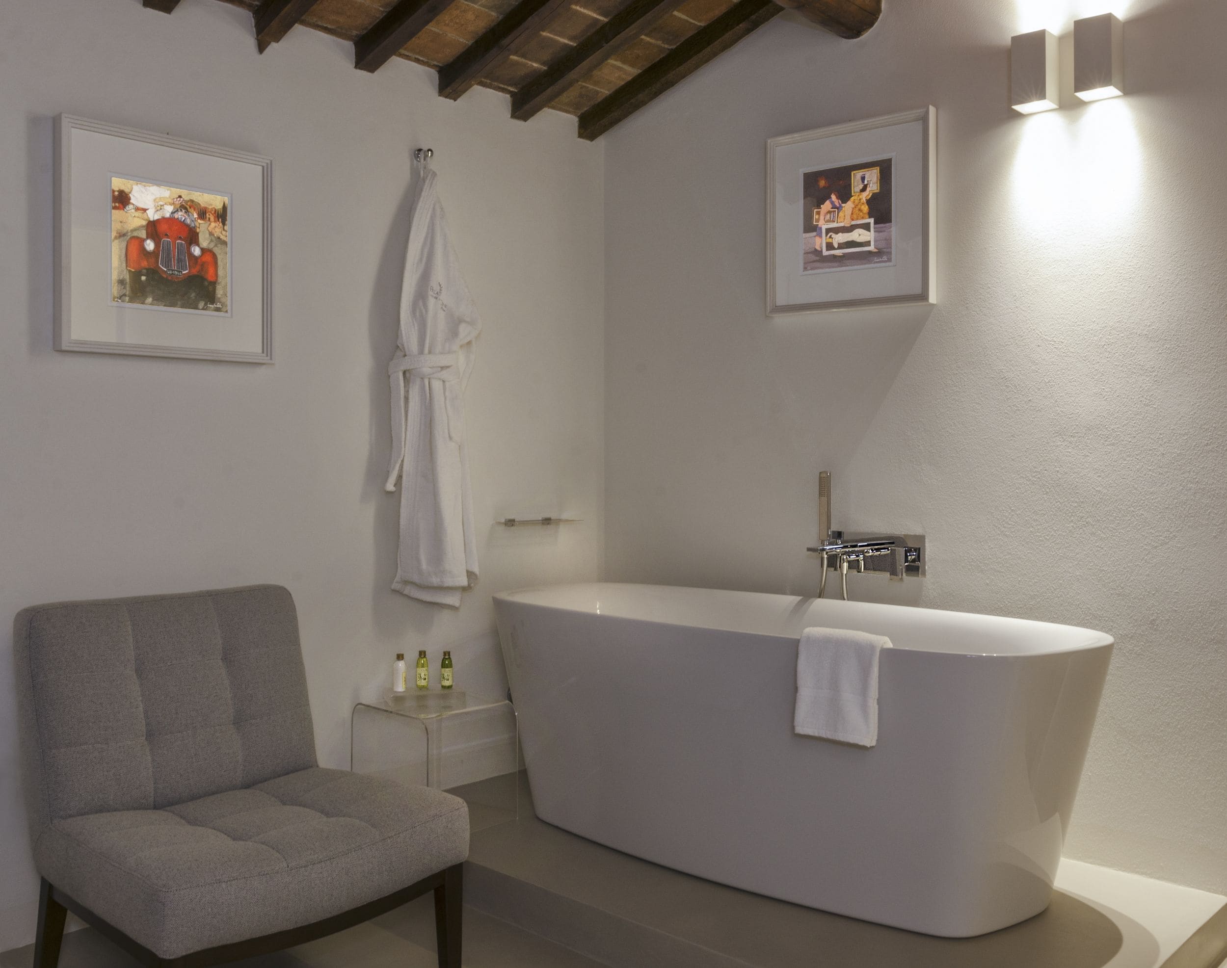 A modern bathroom with a freestanding bathtub, a gray armchair, and two framed pictures on the walls.