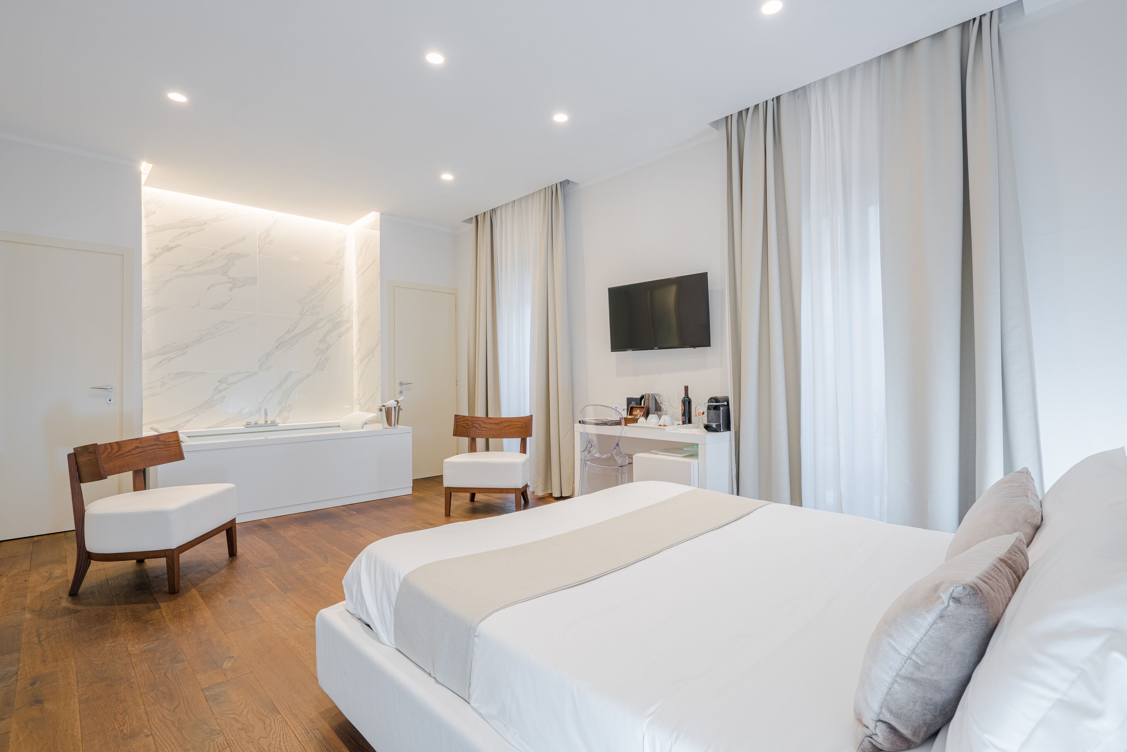 Una habitación de hotel moderna con una cama grande, un escritorio con televisor de pantalla plana, una silla, una mesita y un baño con pared de mármol y bañera.