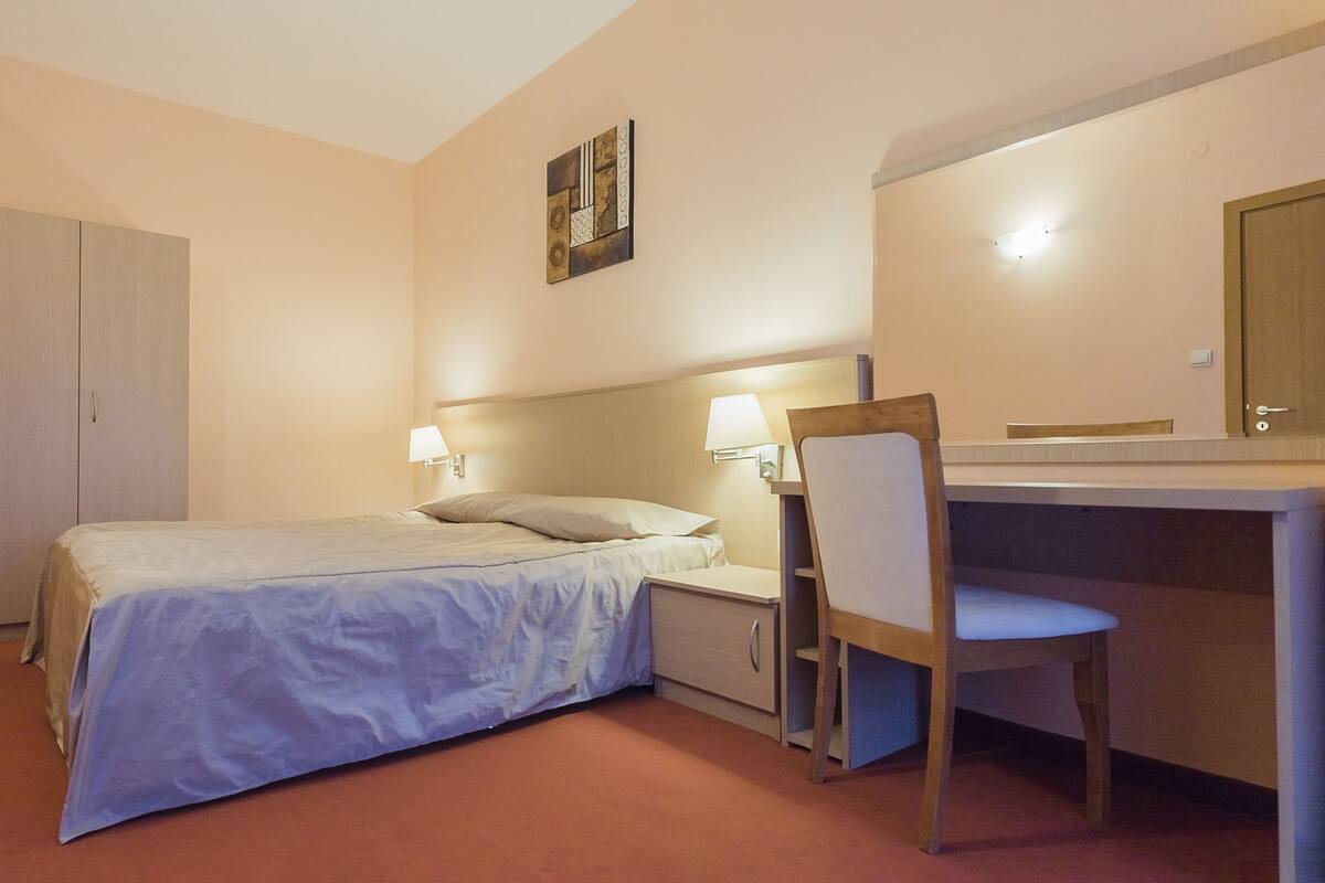 Ett hotellrum med en säng, ett skrivbord, en stol, en garderob och ett konstverk som hänger på väggen.