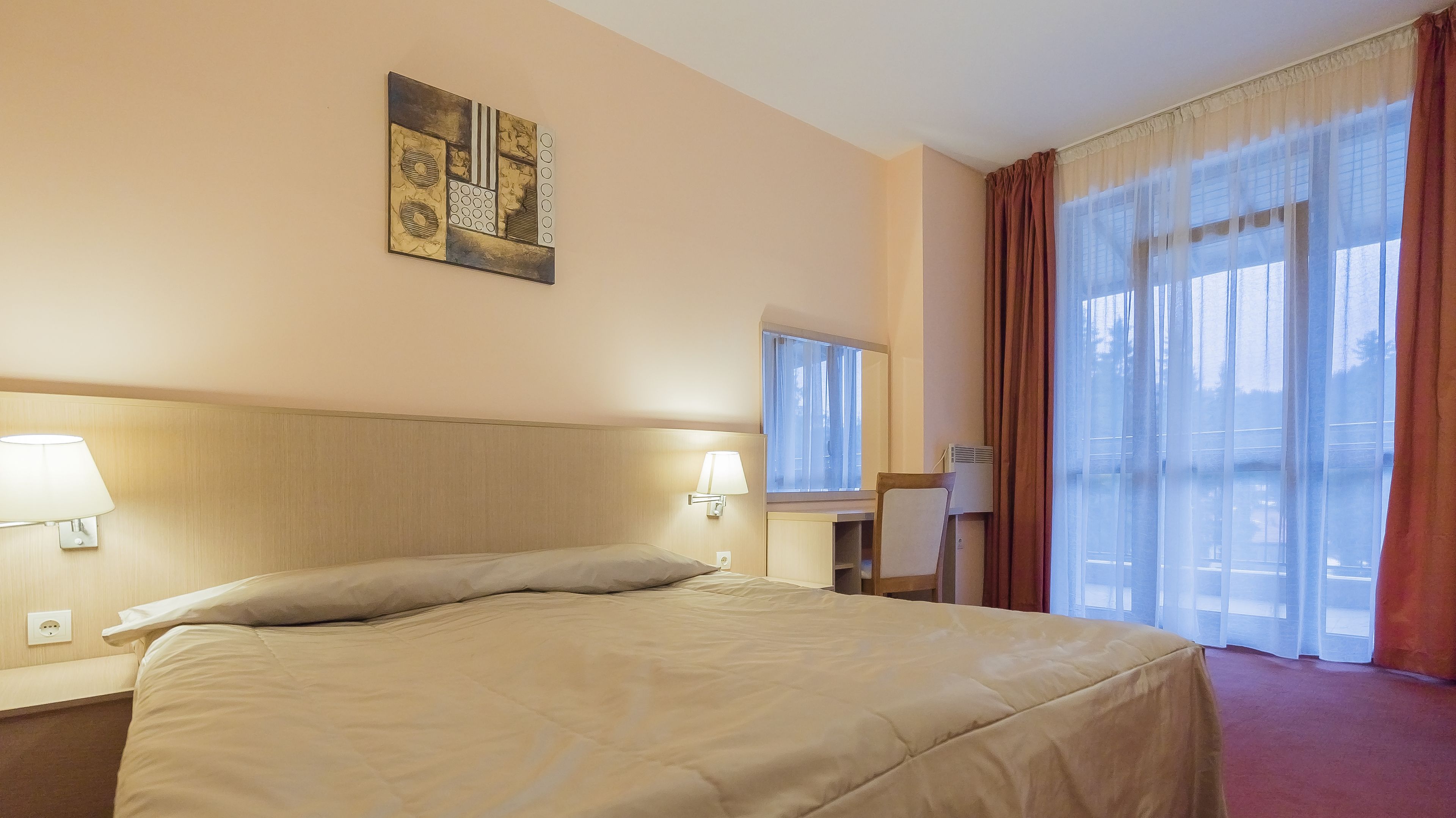 Una habitación de hotel con una cama grande, dos lámparas de noche, un escritorio, una silla, una ventana con cortinas y un cuadro en la pared.