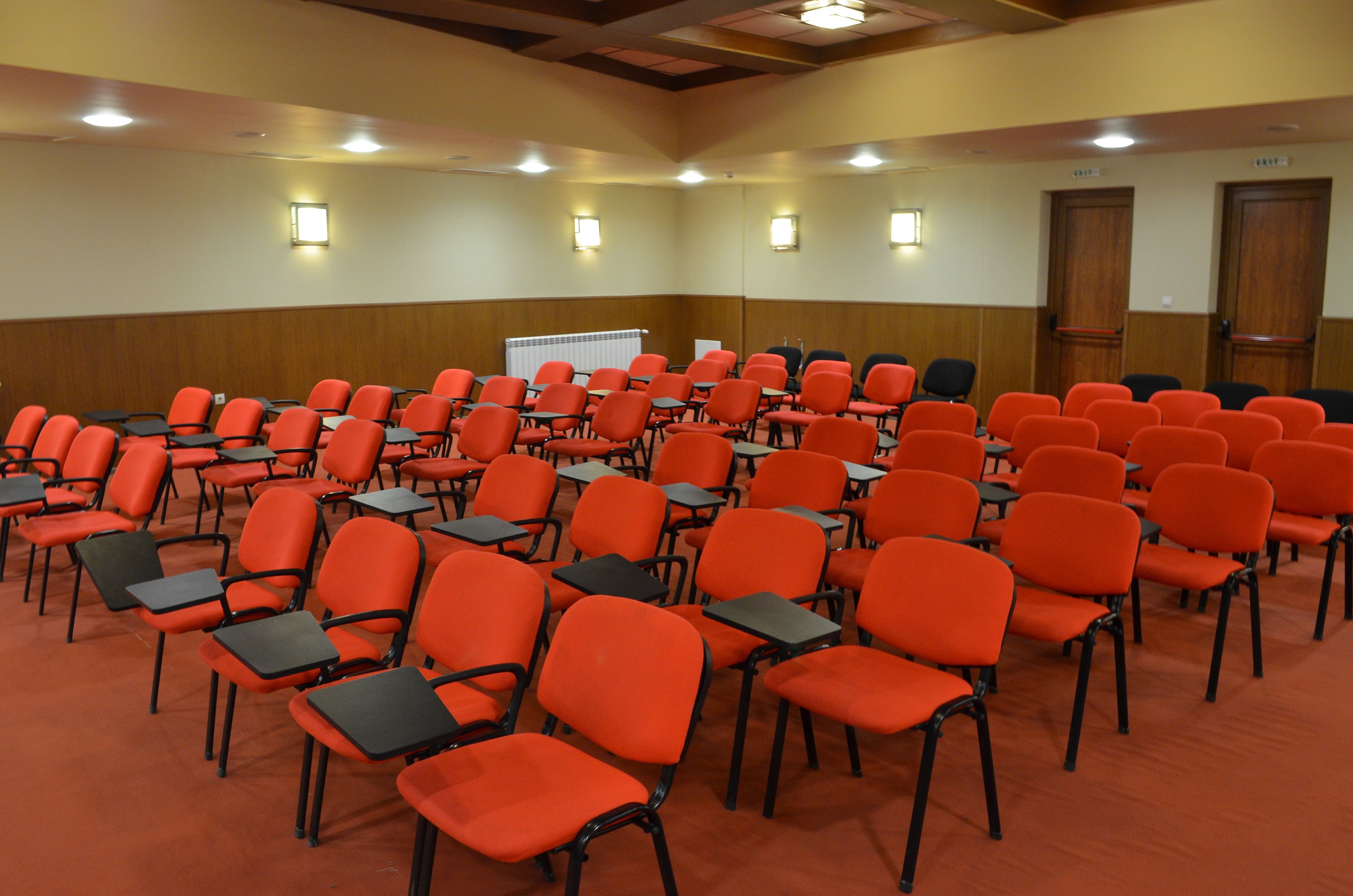 A conference room with rows of orange chairs and black tables, equipped with ceiling lights and wall-mounted lamps.