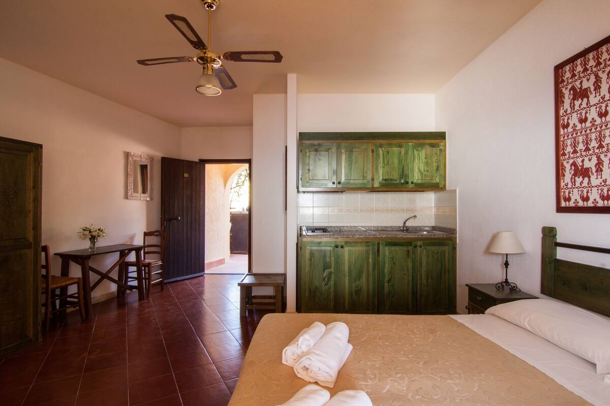 Habitación de hotel con cama, escritorio, silla, mesa con un jarrón de flores, ventilador de techo, armarios verdes, lavamanos y mesita de noche con lámpara.