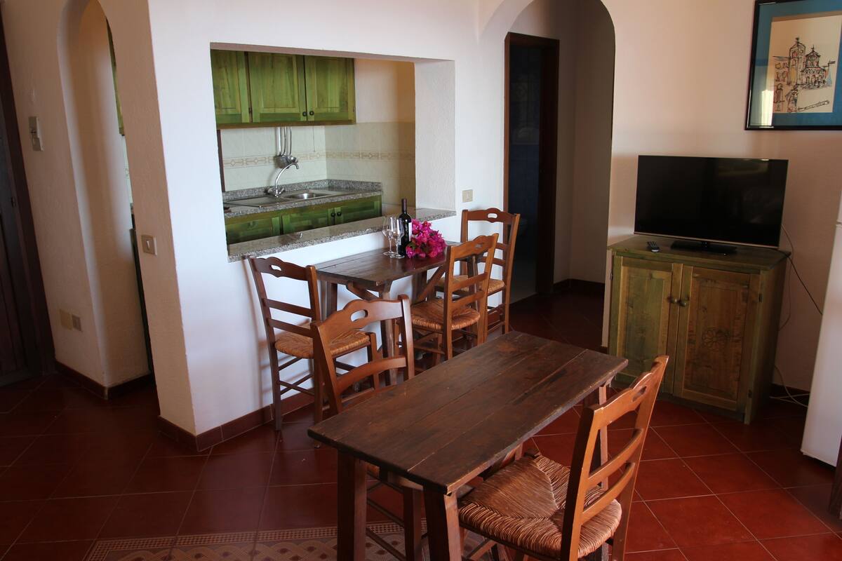 Un comedor con mesa y sillas de madera, una cocina con armarios verdes y un televisor sobre un mueble.