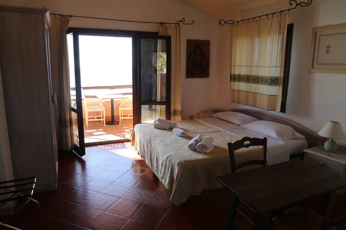 Un dormitorio con cama, una mesa y una silla. Hay una ventana con cortinas y vistas al exterior.
