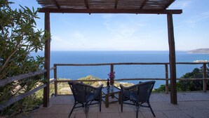 A terrace with a wooden pergola, two wicker chairs, a table with a bottle and flowers, and a view of the sea.
