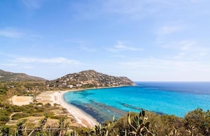 A coastal landscape with a beach, clear blue waters, and a hill with buildings.