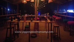 An outdoor bar area with wooden tables and stools, surrounded by wicker lamps.