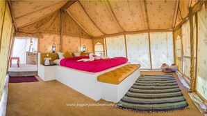 A tented room with a bed, a small table, a lamp, and a view of the outside.