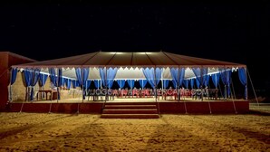 A large, white tent with blue curtains, set on a sandy beach at night.