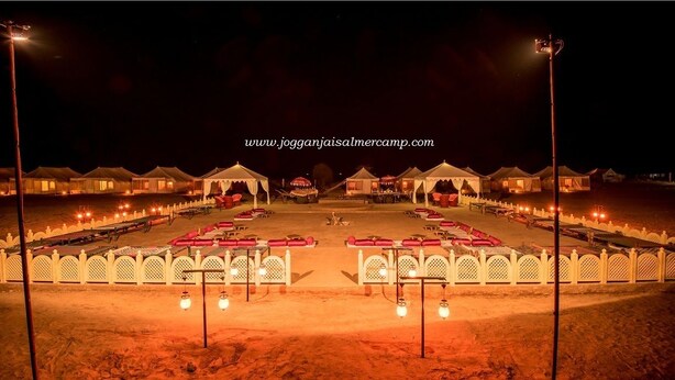 A nighttime view of a camp with multiple tents arranged in rows, each with a small table and chairs.