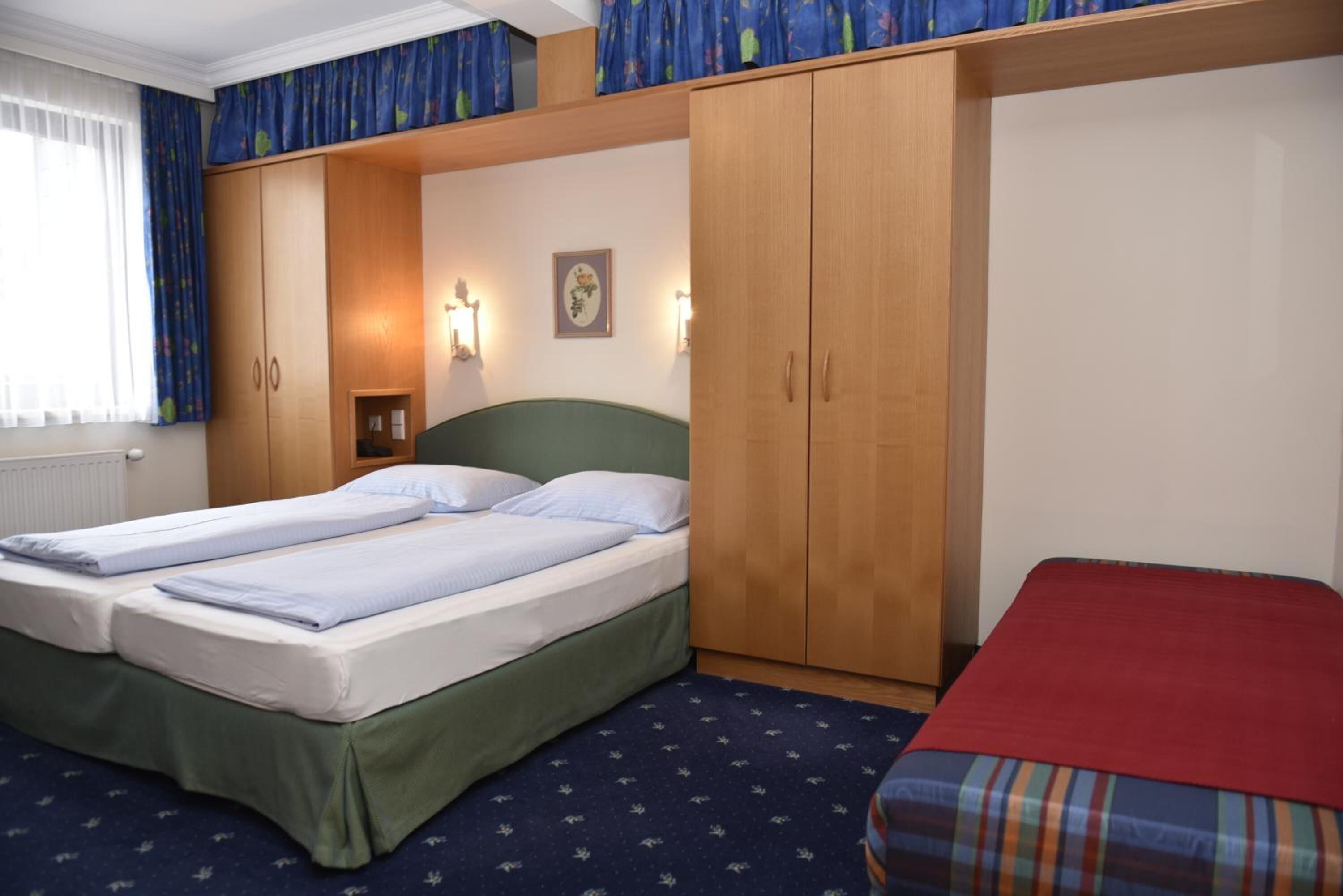 A room with two beds, wooden wardrobe, and a small table with a lamp.