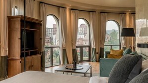 Habitación de hotel con una cama grande, un aparador de madera, una mesita pequeña y un sofá. Cuenta con ventanas amplias con cortinas y vistas a edificios.