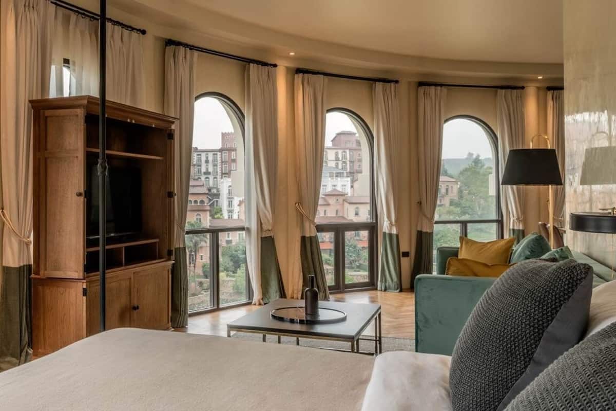 A hotel room with a large bed, a wooden cabinet, a small table, and a sofa. There are large windows with curtains, and a view of buildings outside.