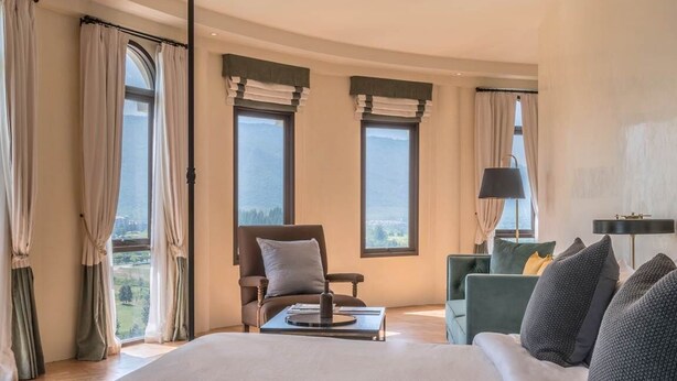 Un dormitorio amplio con una cama grande, una zona de estar con una silla y una mesa, y vistas al exterior a través de amplios ventanales.