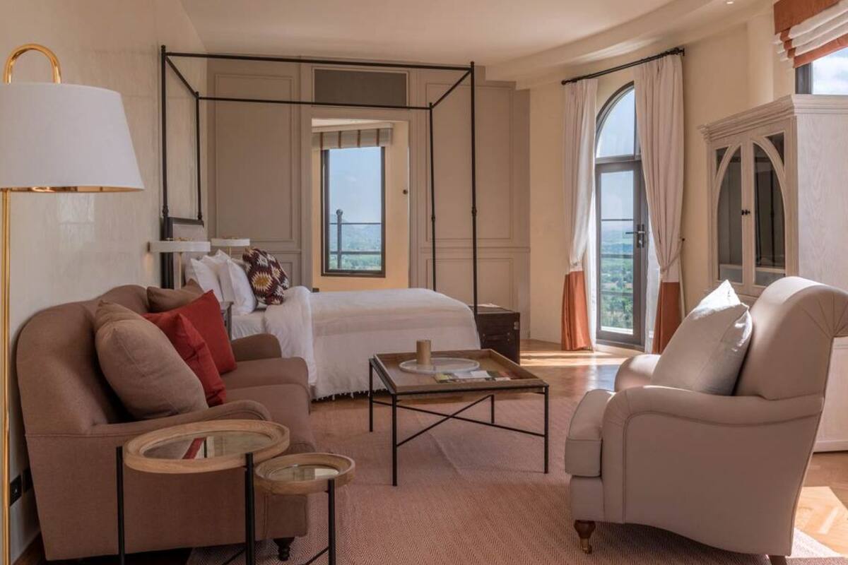 A modern hotel room with a large bed, a sitting area with a sofa and armchairs, a small table, and a large window offering a view.