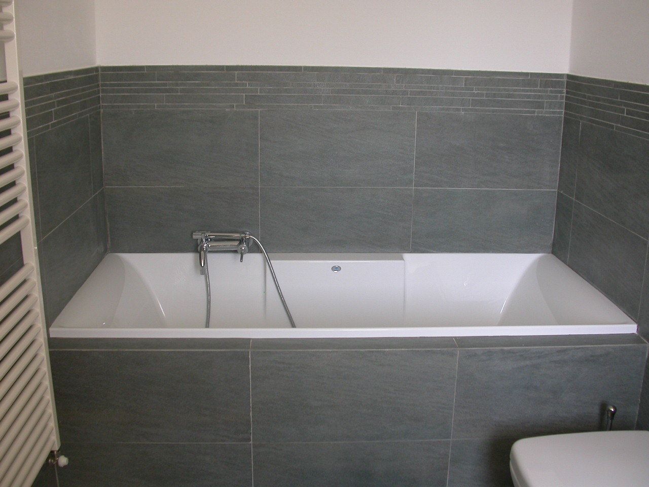 Una bañera rectangular estándar con grifo y ducha, frente a paredes de azulejos grises oscuros.