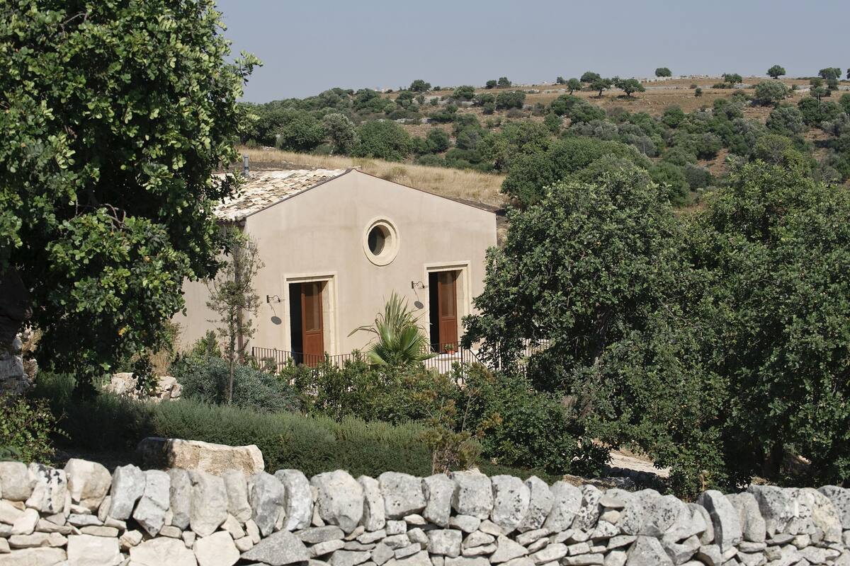 A stone wall in the foreground, a building with two doors and a circular window, surrounded by greenery and trees.