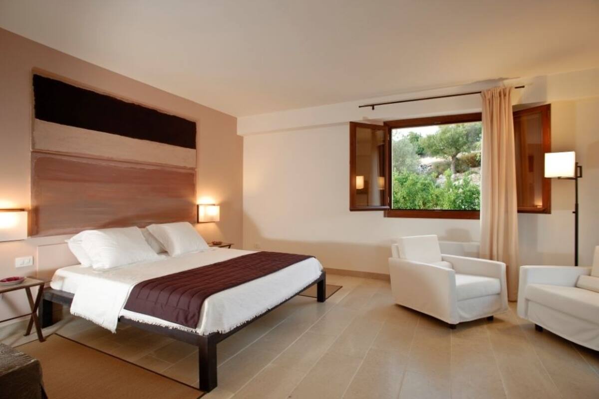 A modern hotel room with a large bed, two white armchairs, a nightstand, and a window with a view of greenery.