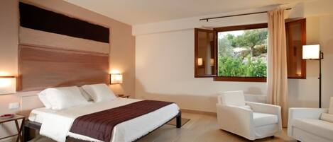 A modern hotel room with a large bed, two white armchairs, a nightstand, and a window with a view of greenery.