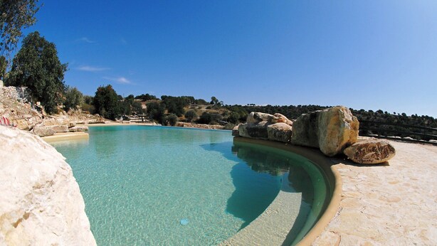 A clear pool with a curved edge, surrounded by large rocks and a stone deck. - Relais Parco Cavalonga (Ragusa)