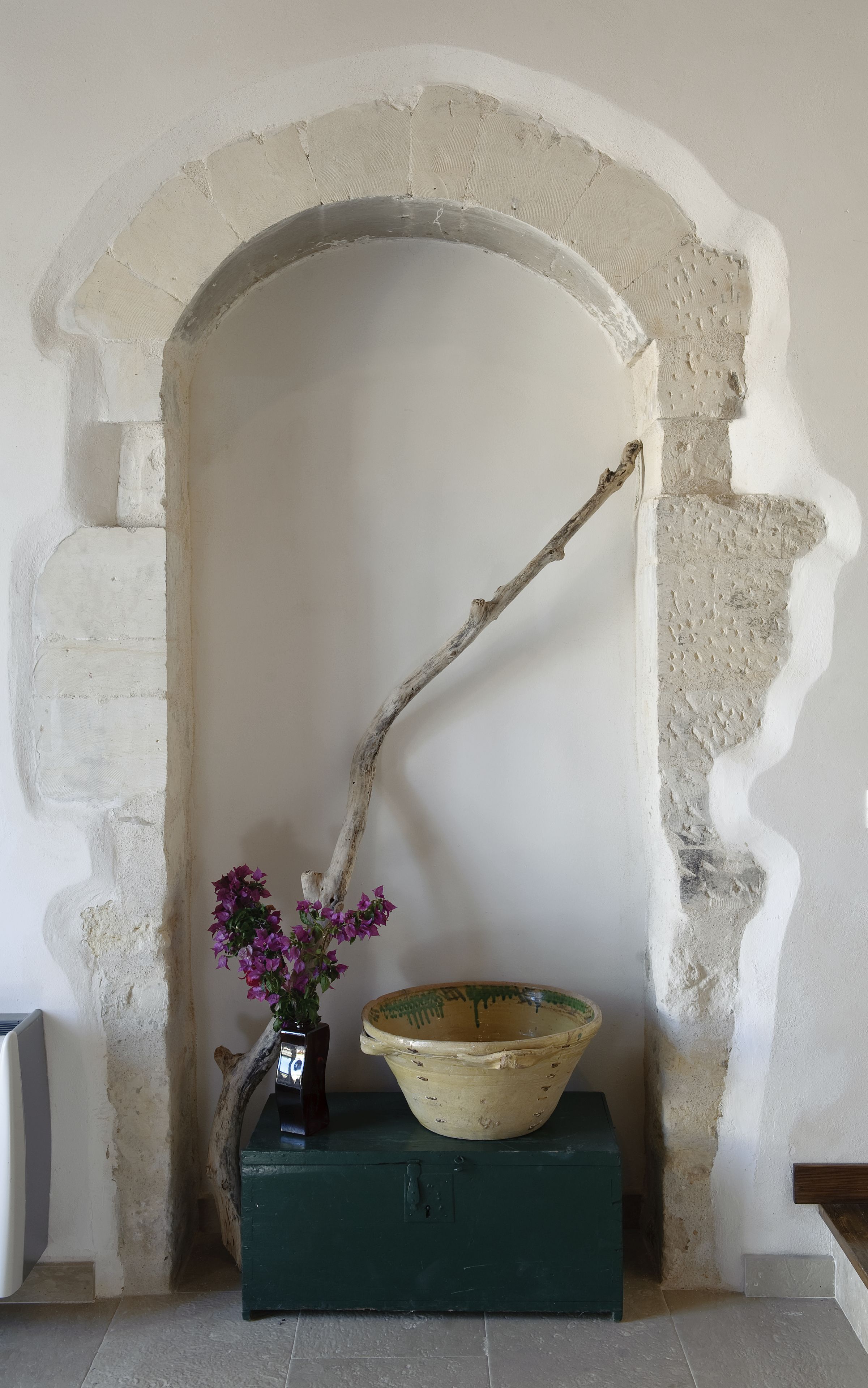 Una habitación con un arco de piedra, un jarrón con flores y un cuenco rústico sobre un caja verde.