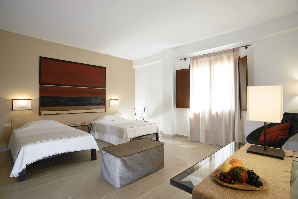 A hotel room with two beds, a sofa, a glass coffee table with fruit, and a painting on the wall.