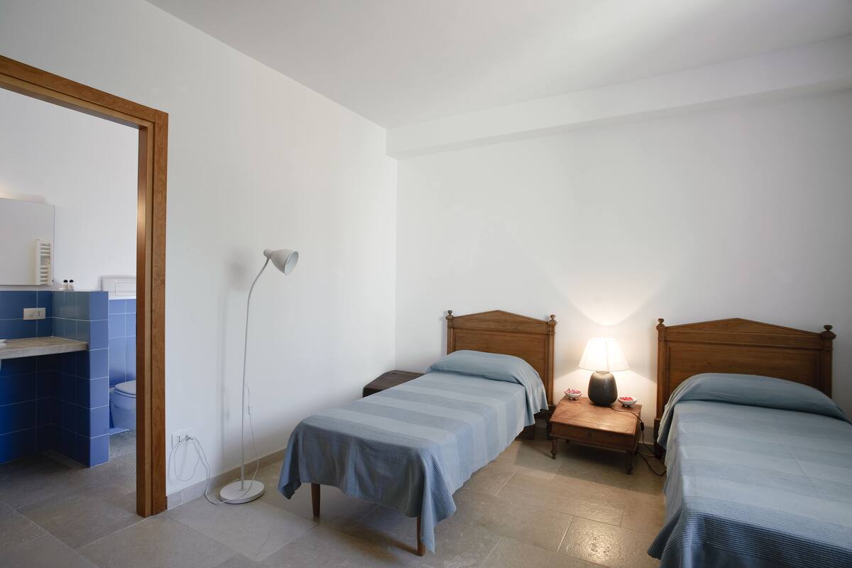 A bedroom with two single beds, a bedside table with a lamp, and a small bedside table with a bowl of fruit.