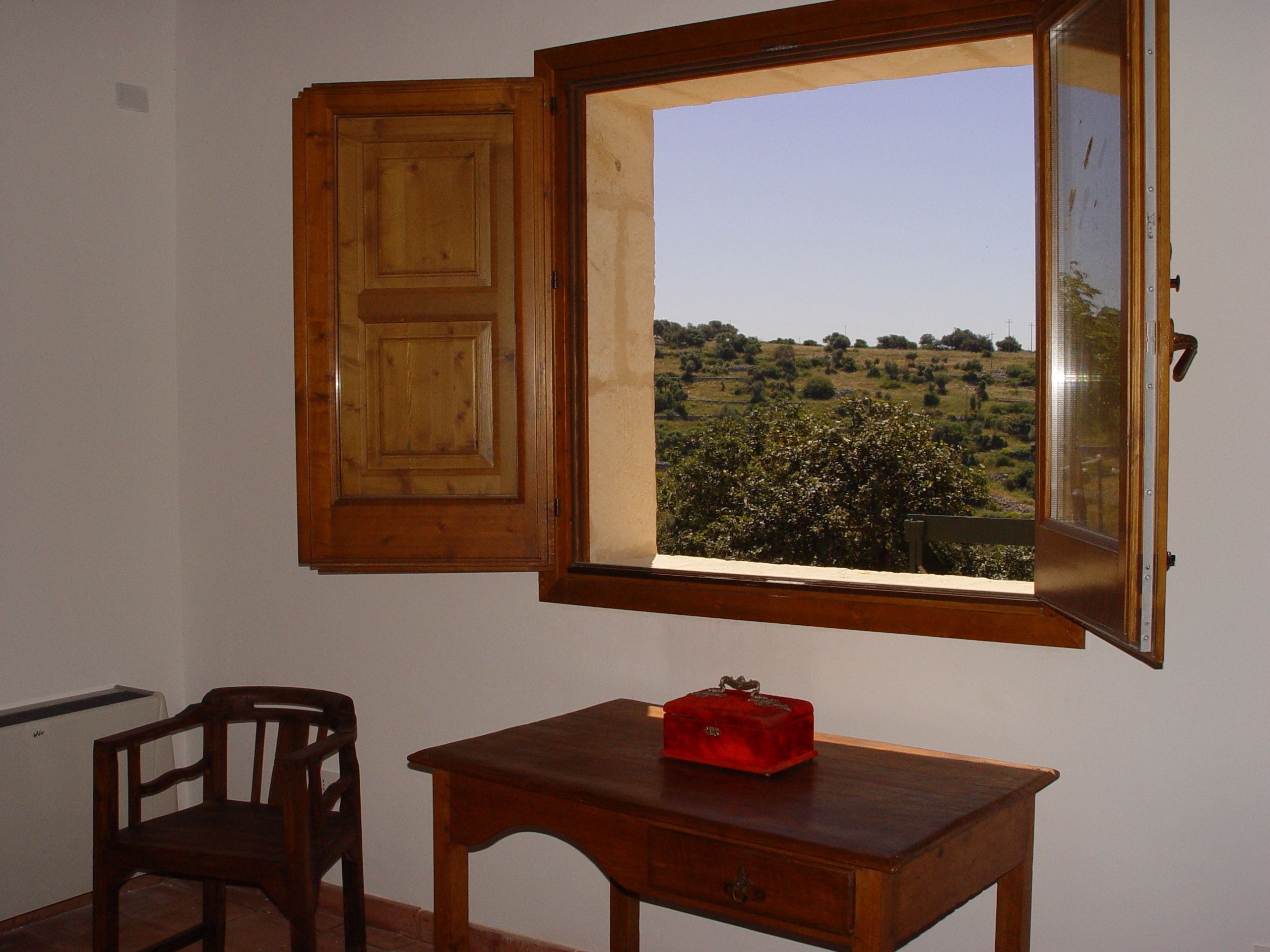 A room with a wooden cabinet, a chair, a small table with a red box, and a window with a view of a green landscape.
