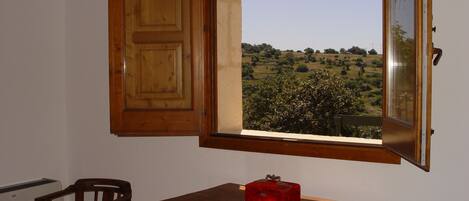 Una habitación con un armario de madera, una silla, una mesita con una caja roja y una ventana con vistas a un paisaje verde.