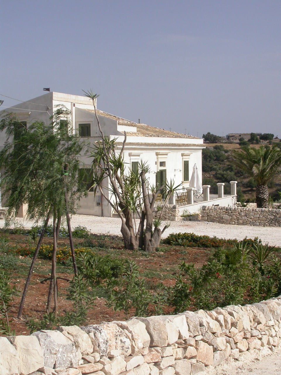 A white building with a stone wall in front, surrounded by greenery and a clear sky.