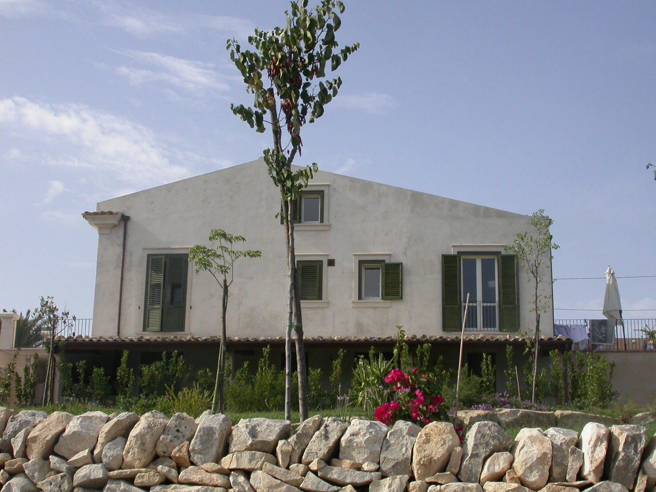 Un bâtiment à deux étages, doté d’un toit plat, avec un mur de pierre au premier plan et un petit arbre au centre.
