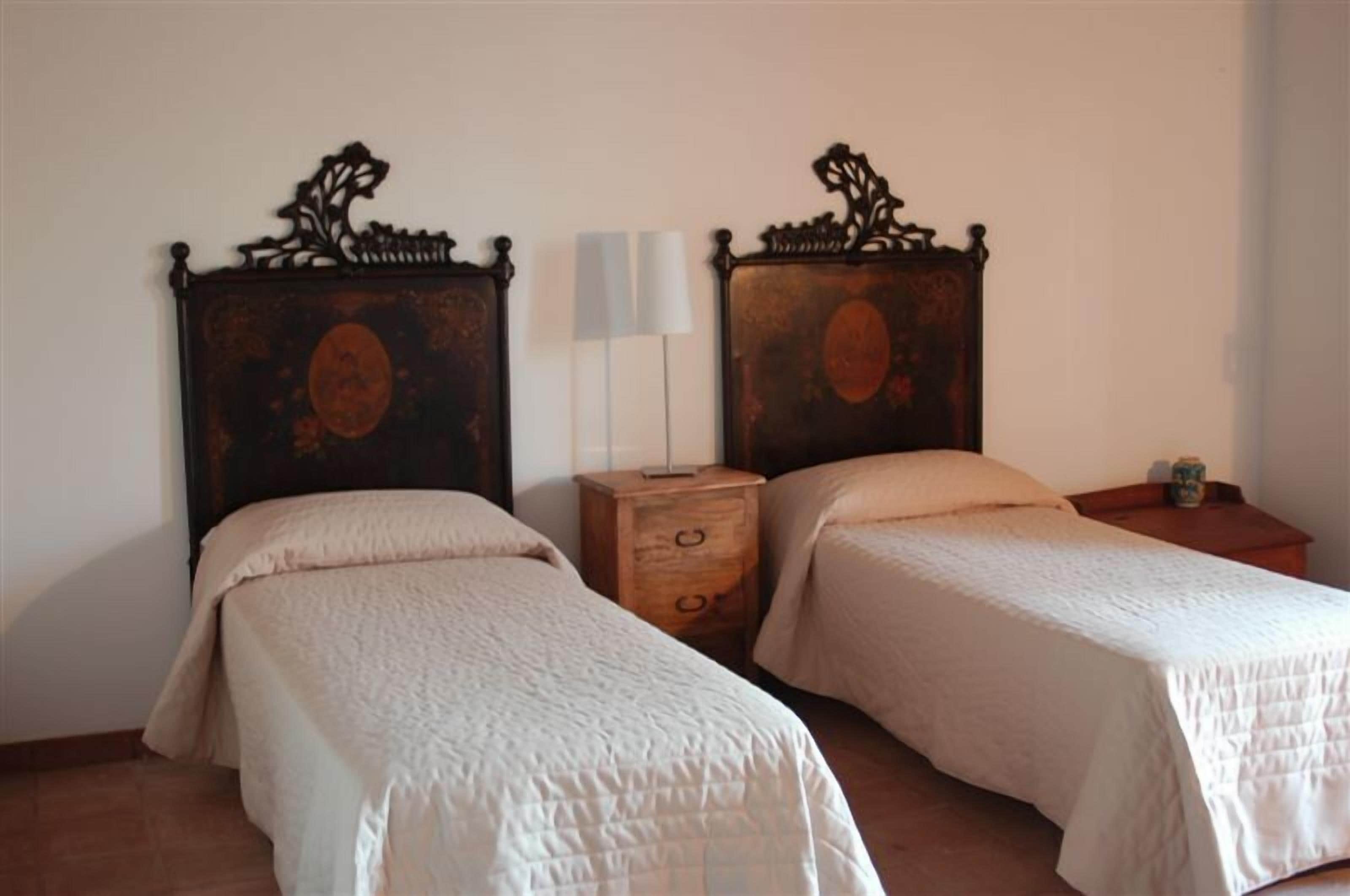 A room with two beds, each with a wooden headboard and a white bedspread. There is a nightstand with a lamp between the beds.