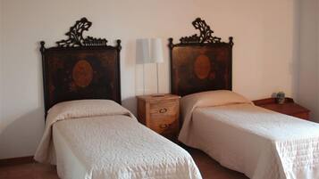 A room with two beds, each with a wooden headboard and a white bedspread. There is a nightstand with a lamp between the beds.