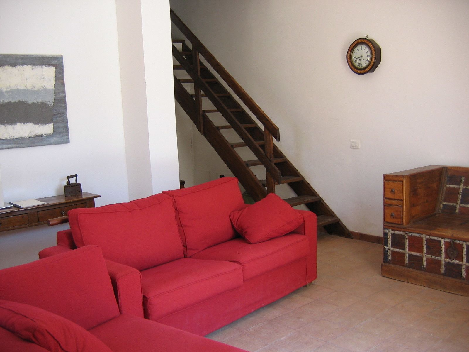 A living room with a red sofa, a wooden chest of drawers, a staircase, and a wall clock.