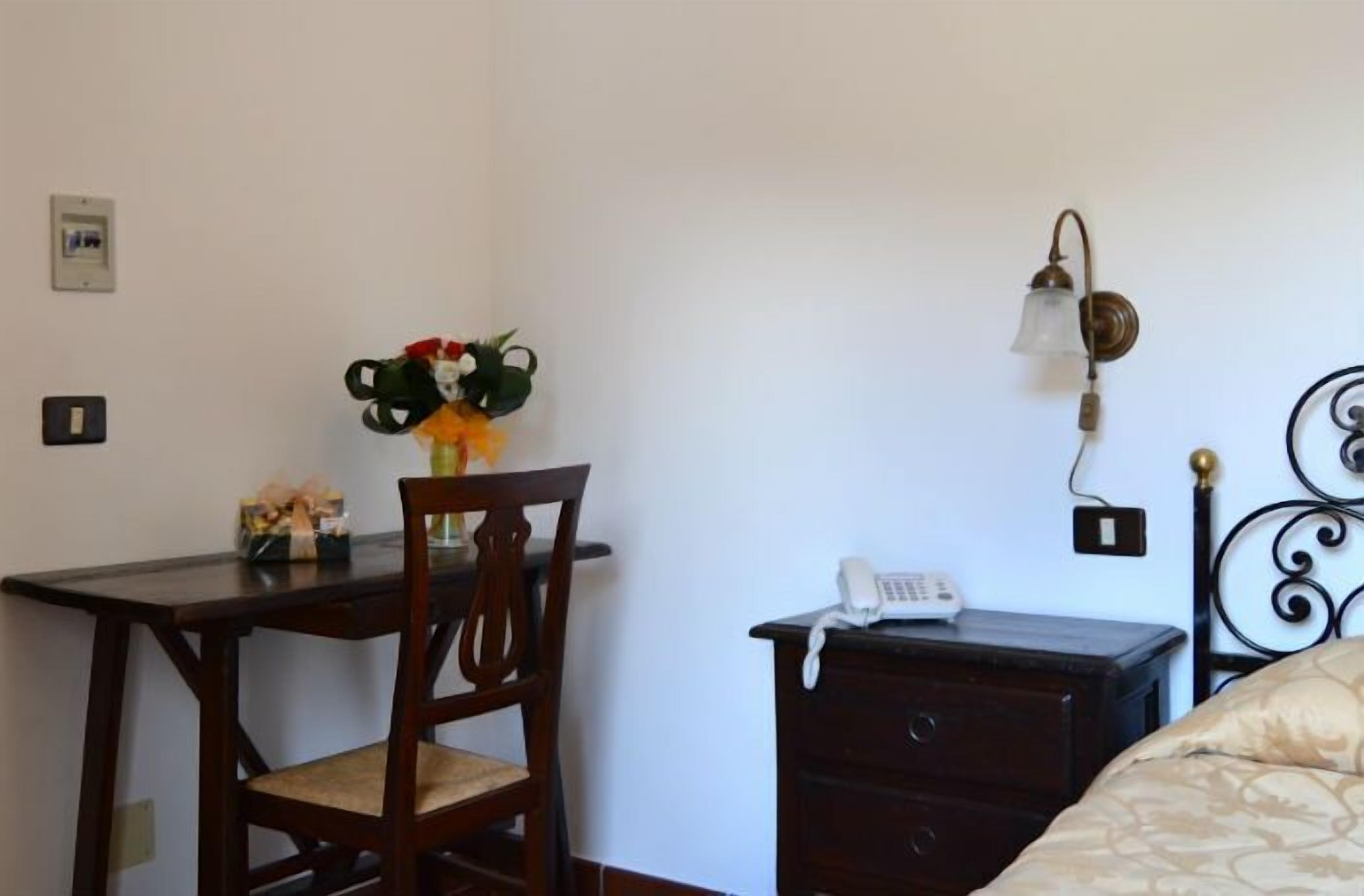 A room with a wooden desk, a chair, a bedside table with a phone, a wall-mounted light fixture, and a bed with a patterned bedspread.
