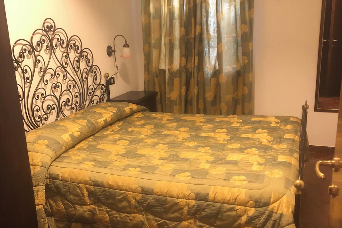 Un dormitorio con cama, una mesita de noche, una lámpara de pared, una cortina y una puerta con picaporte.