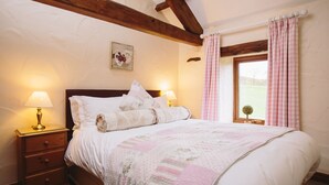 A neatly made bed with a quilt, two bedside lamps, a wooden nightstand with drawers, and a window with pink curtains. - Garth Cottage (Brampton)