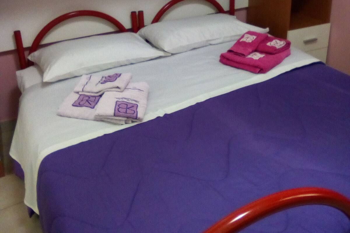 A single bed with a purple bedspread, two pillows, and a set of pink towels. There is a wooden headboard and a white wardrobe above the bed.