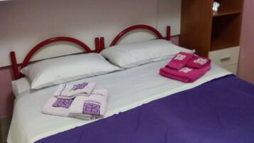 Una cama individual con una colcha morada, dos almohadas y un juego de toallas rosas. Tiene un cabecero de madera y un armario blanco sobre la cama.