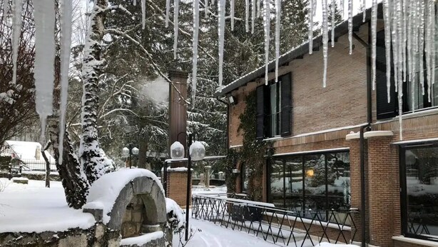 A snow-covered courtyard with a brick building, a stone fountain, and a snow-covered tree. - LH Hotel il Picchio (Pescasseroli)