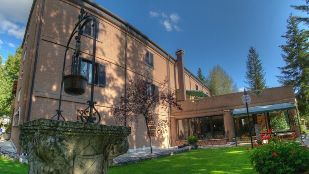 A brick building with a covered entrance, a stone fountain, and a well-maintained lawn with red flowers. - LH Hotel il Picchio (Pescasseroli)