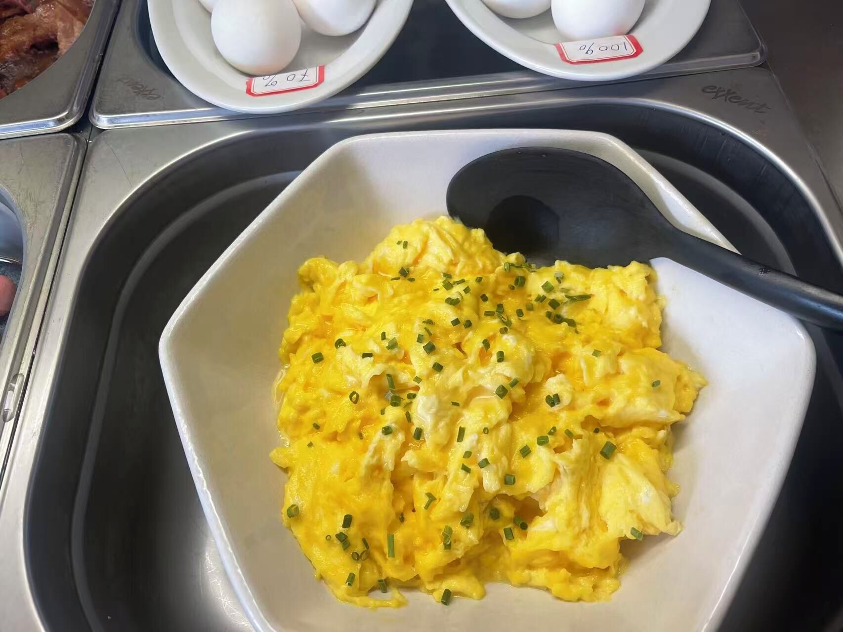 A bowl of scrambled eggs garnished with chives, placed on a tray with a black spoon.