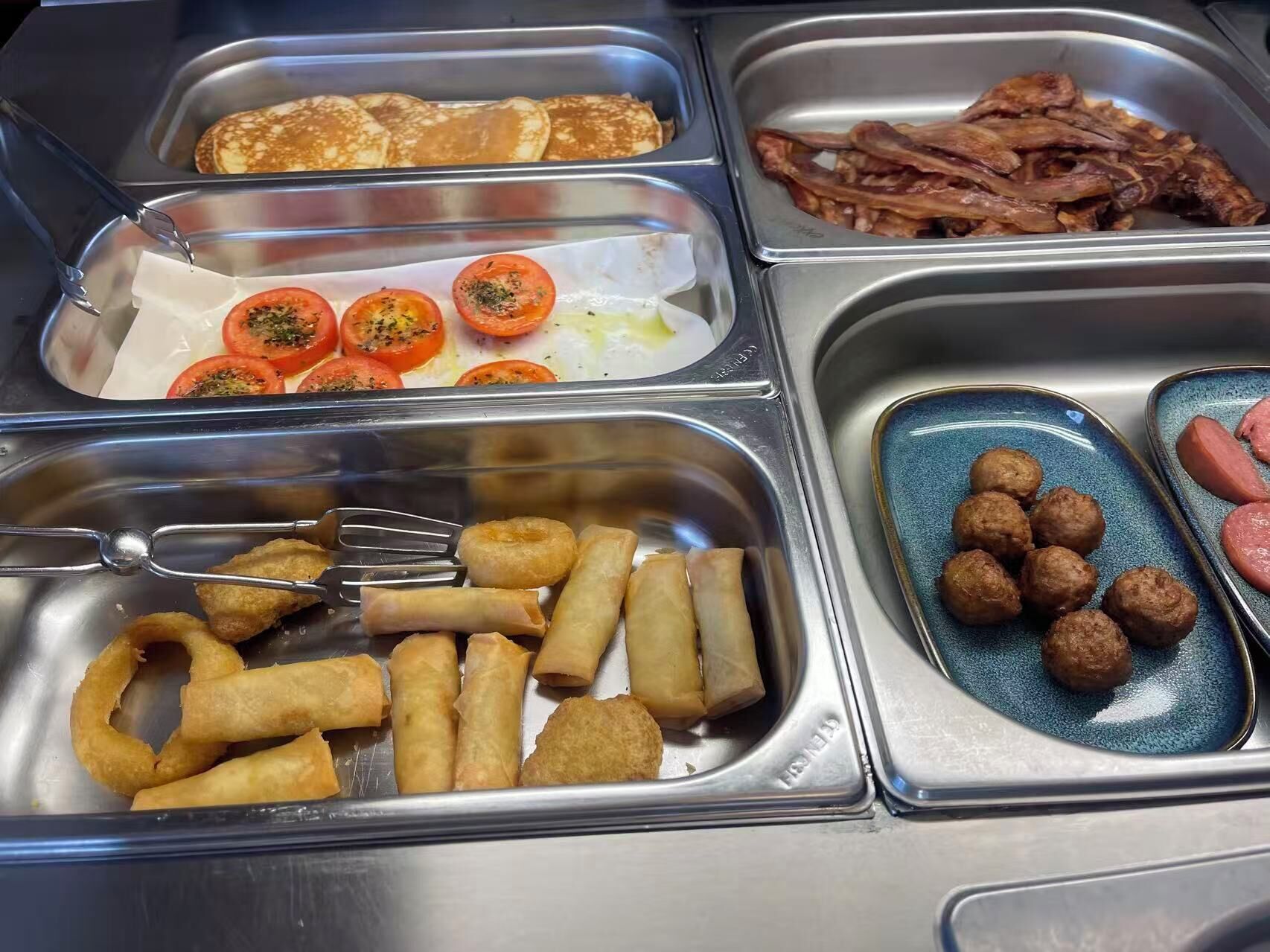 Ein reichhaltiges Buffet mit verschiedenen Speisen, darunter Pfannkuchen, Speck, Tomaten, Frühlingsrollen und Frikadellen.
