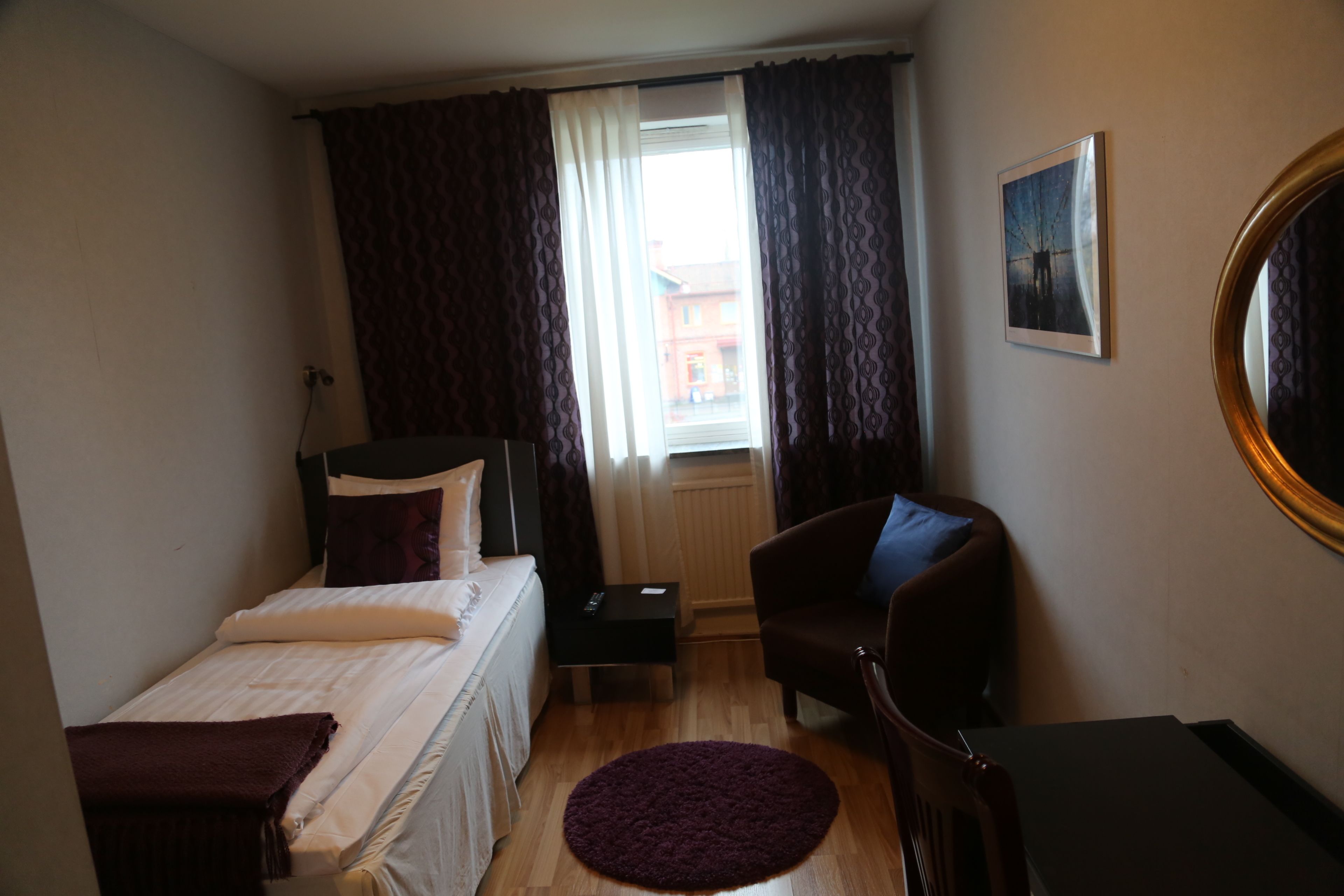 Un dormitorio con cama, una silla, una mesita, una ventana con cortinas y un cuadro en la pared.