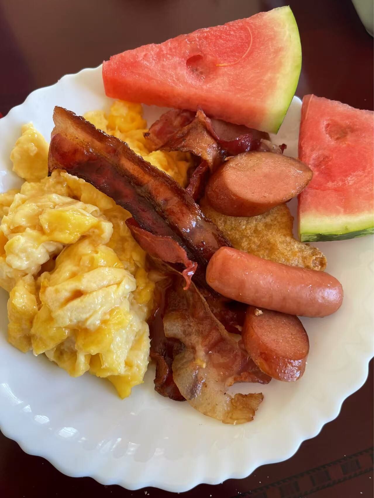 A plate with scrambled eggs, bacon, sausages, and watermelon slices.
