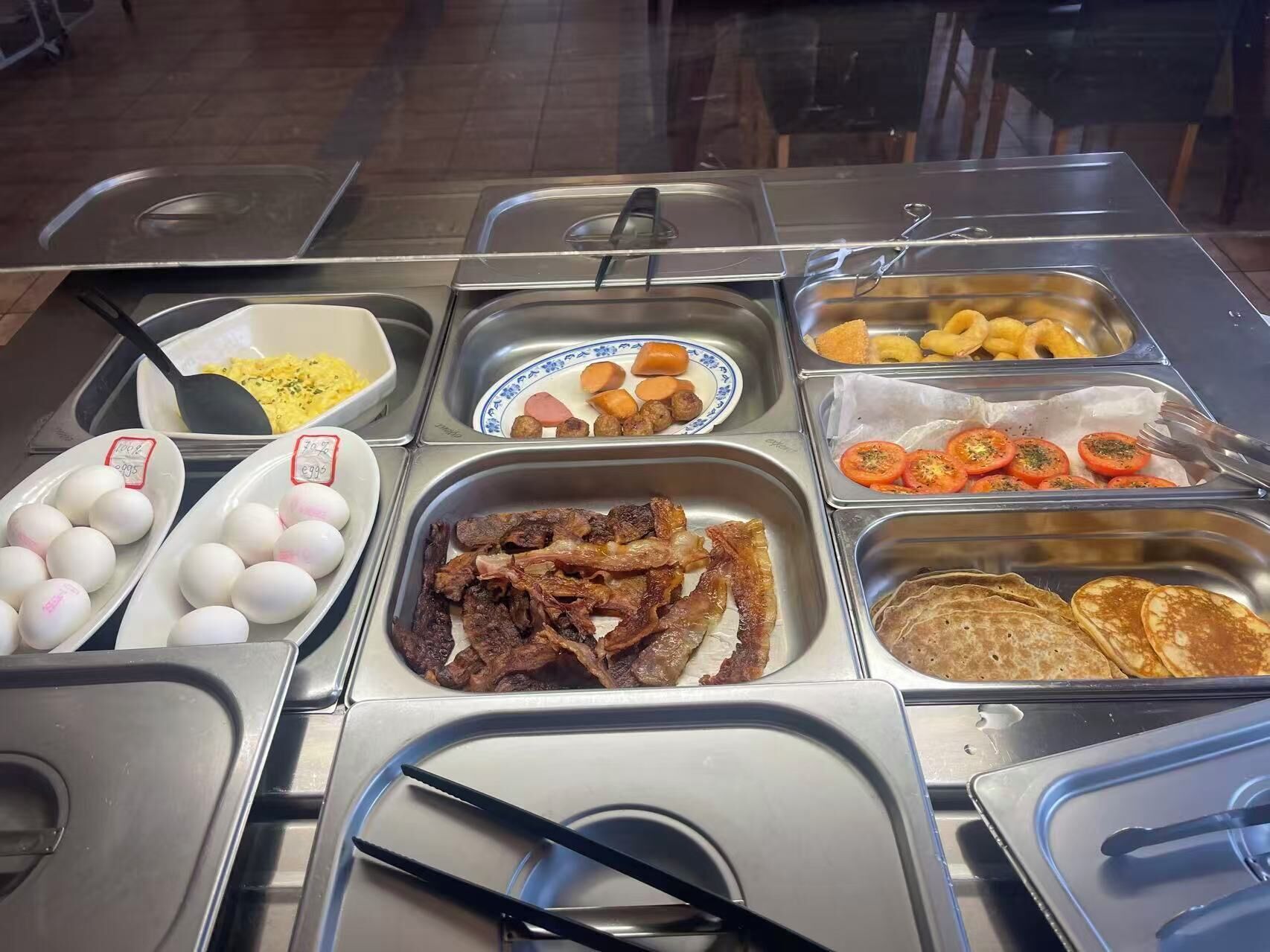 Un bufé con diversas opciones de comida, incluyendo huevos, tocino, tomates y panqueques.
