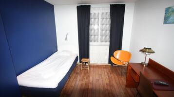 Una camera d'albergo con un letto singolo, un tavolino in legno, una sedia, una scrivania con una lampada e una finestra con tende.