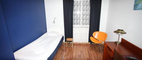 Quarto de hotel com uma cama de solteiro, uma pequena mesa de cabeceira de madeira, uma cadeira, uma escrivaninha com luminária e uma janela com cortinas.